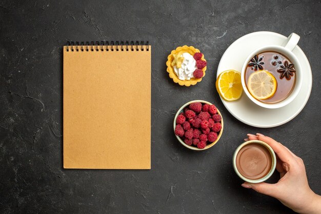 ノートブックの俯瞰図と暗い背景にチョコレートラズベリー蜂蜜を添えてレモンと紅茶のカップ