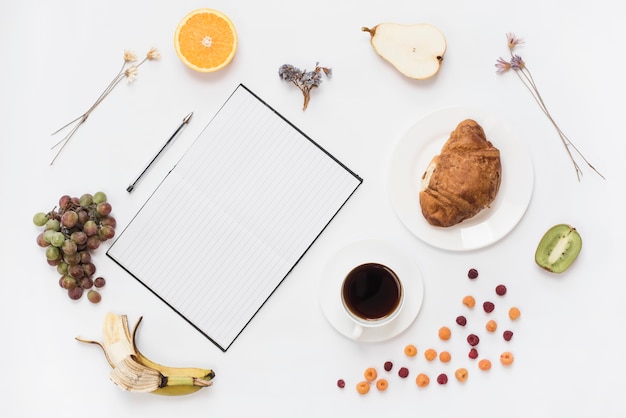 An overhead view of notebook; pen with croissant and healthy fruits isolated on white background