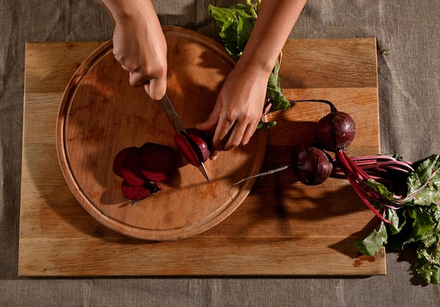 木の板でビートの根を切ったり、切り刻んだり、スライスしたりする女性の手の俯瞰図。新鮮な野菜を切るナイフで手のクローズアップ。 Premium写真