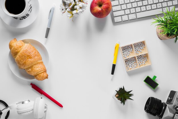 An overhead view of an office stationeries with baked croissant and apple on white desk