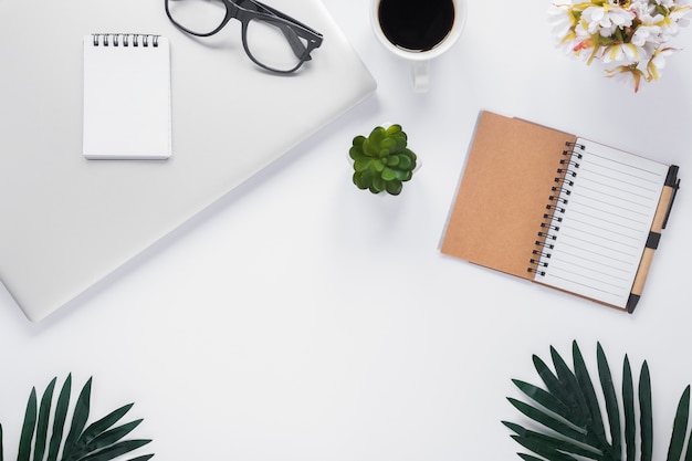 An overhead view of office stationery with laptop; coffee cup; flower vase and leaves on white backdrop