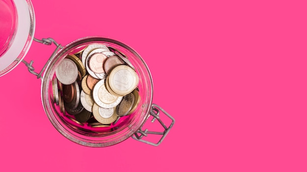 Верхний вид открытой стеклянной банки со многими монетами на розовом фоне