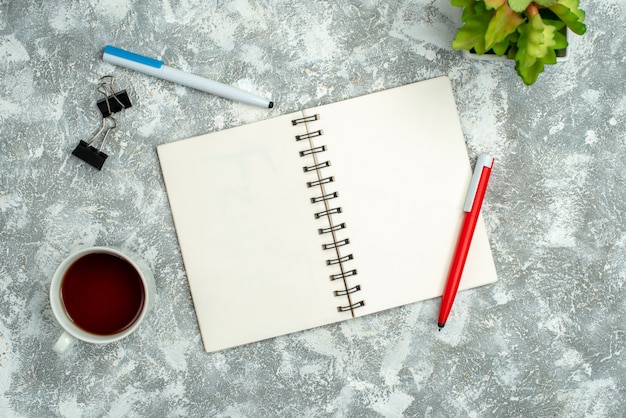 Overhead view of open spiral notebook with two pens and a cup of tea flower pot on gray background