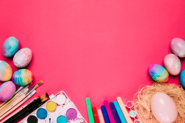 An overhead view of painted watercolor easter eggs; paint brushes; felt tip pen and easter eggs on pink backdrop