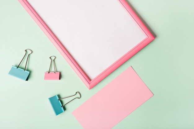 An overhead view of paper clips; blank pink paper and white frame on pastel colored background