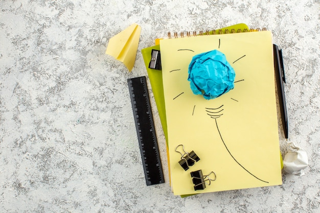 Overhead view of paper light bulb on closed spiral notebooks and office appliances on the left side on white surface