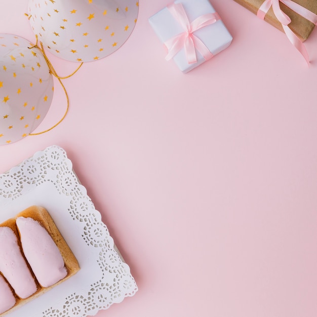 An overhead view of party hat; gift box; and iced buns on pink background