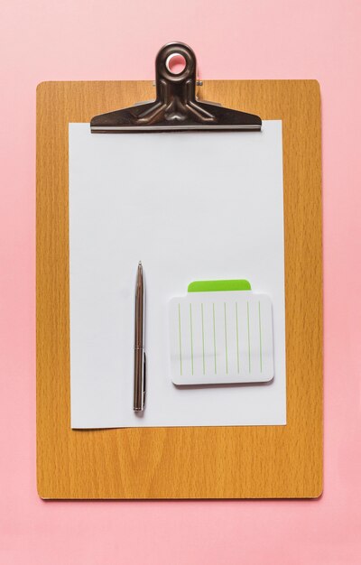 An overhead view of pen and notepad on blank paper over wooden clipboard against pink background