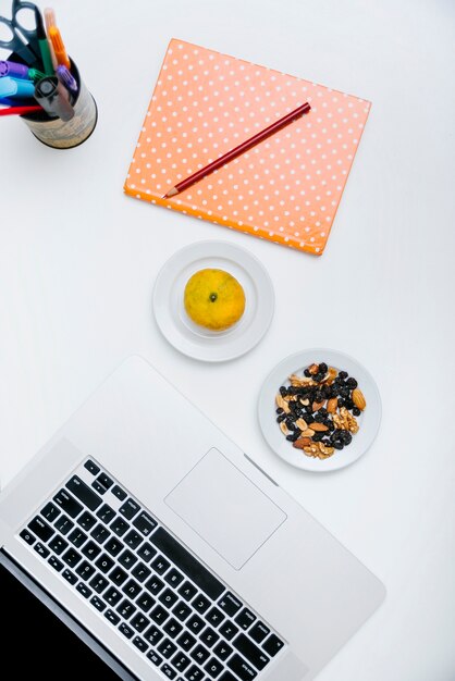 Overhead view of pencil; notebook; citrus fruit; nut food and laptop on white surface