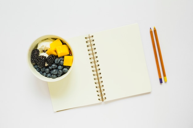 An overhead view of pencils and blank spiral notebook with healthy fruits in cup