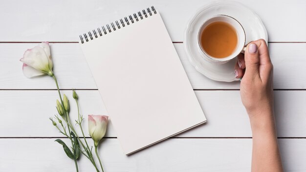 An overhead view of a person holding cup of tea with blank notepad and eustoma flowers