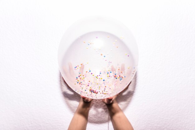 An overhead view of a person's hand holding white balloon with colorful sprinkle on white textured backdrop