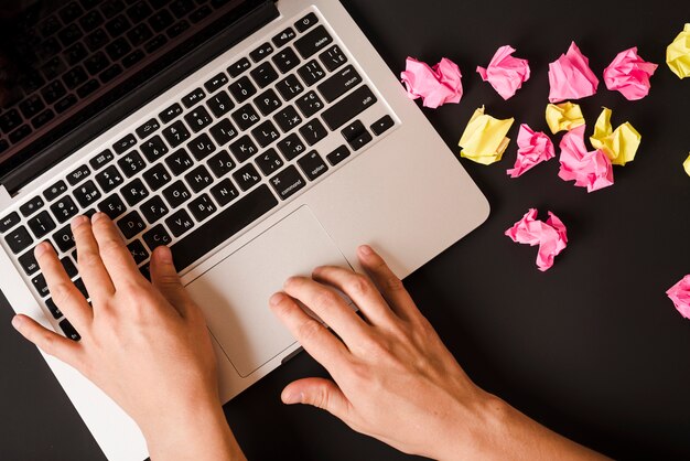 An overhead view of a person's hand typing on laptop with crumpled papers on black background