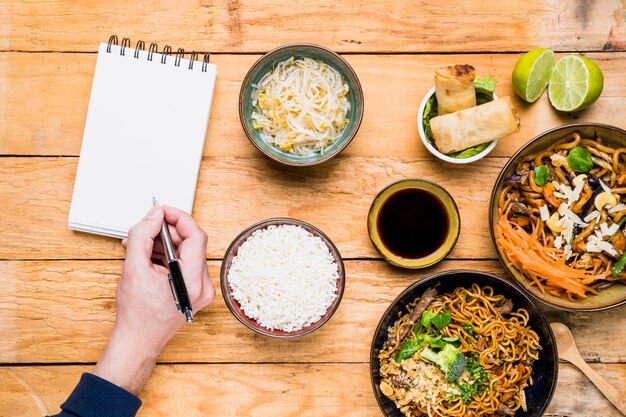 An overhead view of a person writing on pen with spiral notepad with thai traditional food