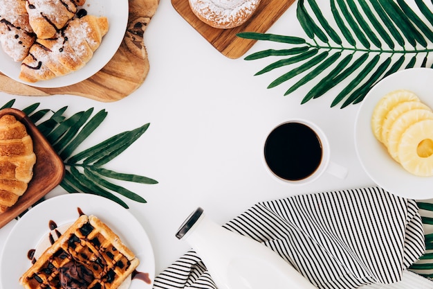 Free photo an overhead view of pineapple slices; baked croissant; waffles; buns; tortillas; milk bottle and coffee on white backdrop