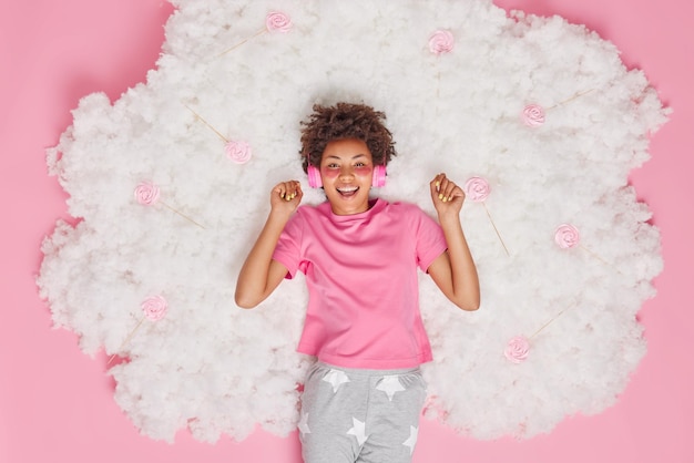 Overhead view of positive curly haired woman enjoys calm music via headphones dressed in pajama applies beauty patches under eyes poses on white fluffy cloud with candies around Rest concept