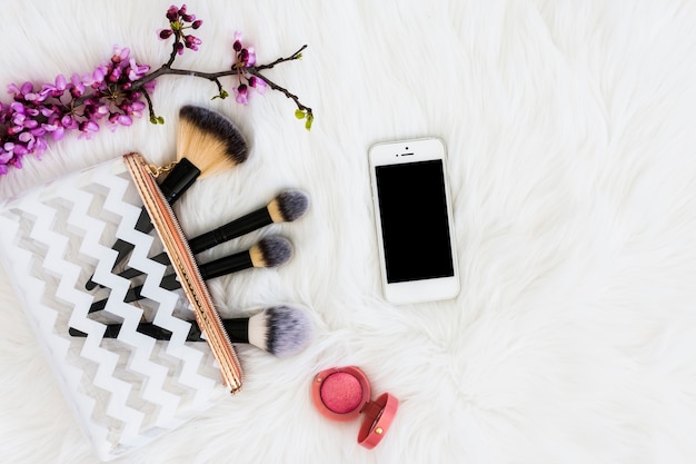 An overhead view of purple twig with makeup brushes; mobile phone and pink compact face powder on white fur