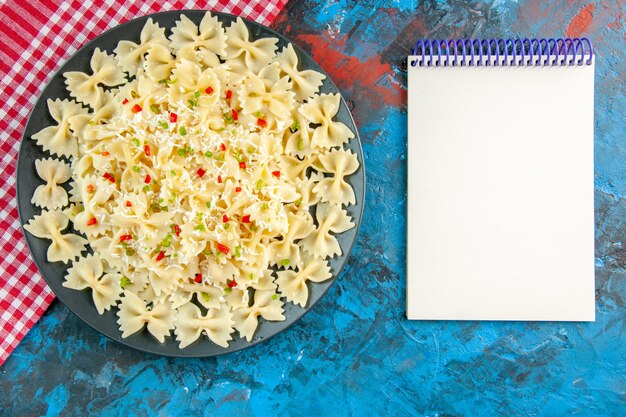 Overhead view of raw Italian farfalle pasta on red stripped towel and spiral notebook on blue background