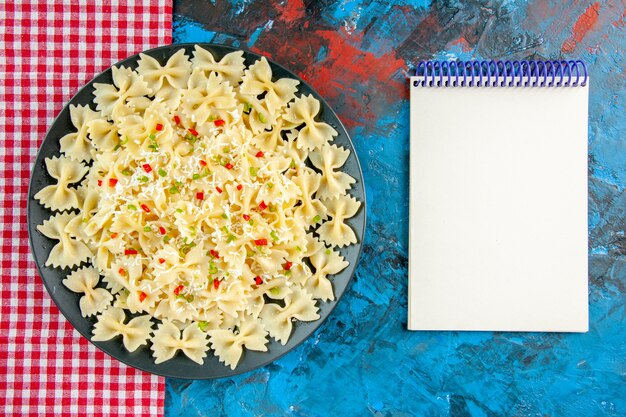 赤いストリップタオルと青いテーブルの上のスパイラルノートに野菜と生のイタリアのファルファッレパスタの俯瞰図