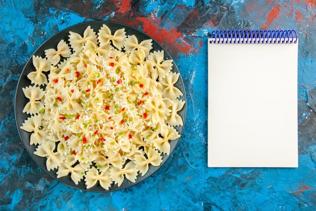 Вид сверху сырой итальянской пасты фарфалле с овощами и спиральной записной книжкой на синем фоне