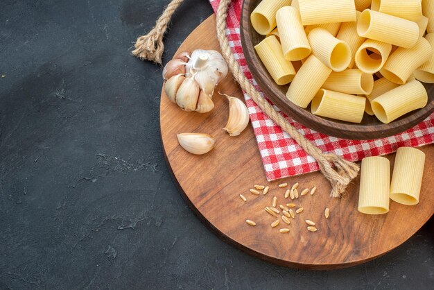 Overhead view of raw pastas inside and outside a brown bowl on red stripped towel garlics rice on round wooden board rope on black background