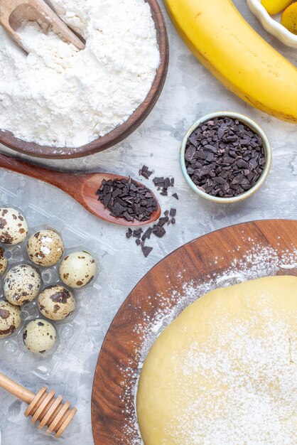 Overhead view of raw pastry flour on round board wooden spoon chocolates eggs banana on stained white background
