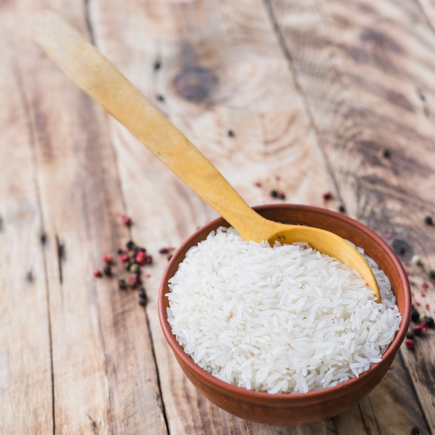 Overhead view of raw white rice bowl with spoon near black pepper spread on wooden table