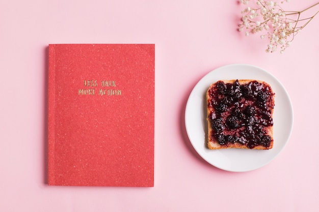 An overhead view of red book; toast with berry jam and baby's breath flowers over pink background