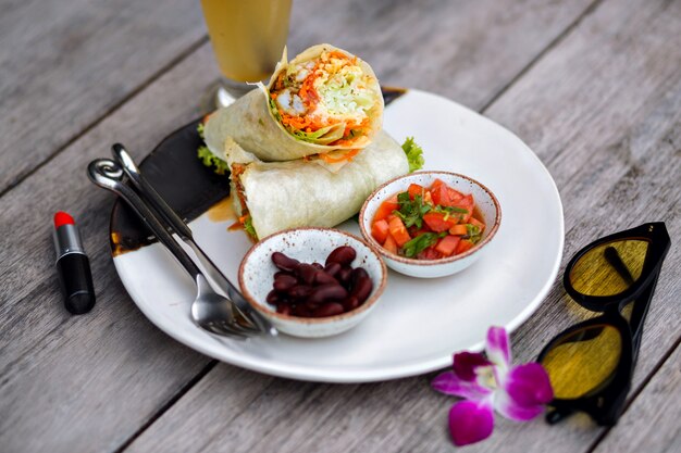 Overhead view of red lipstick, food and purple flower on wooden table. Photo of big plate with tasty salad and beans standing beside glass of smoothie.