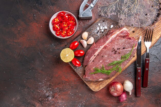 Вид сверху красного мяса на деревянной разделочной доске и чеснока, зеленого лимона, лука, вилки и ножа на темном фоне
