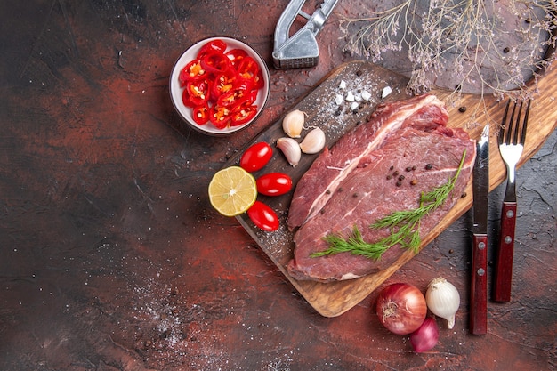 Overhead view of red meat on wooden cutting board and garlic green lemon onion fork and knife on dark background