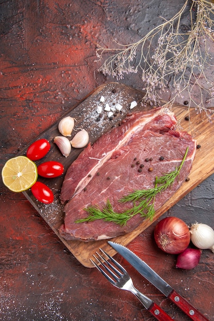 Overhead view of red meat on wooden cutting board and garlic green lemon onion fork and knife on dark background