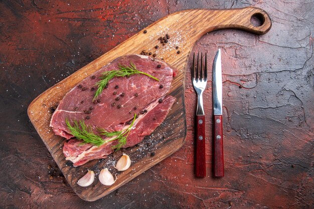 Overhead view of red meat on wooden cutting board and garlic green pepper fork and knife on dark background