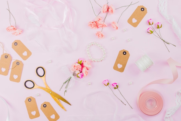 An overhead view of ribbon; artificial rose; pearls; ribbon; tag and scissor on pink background