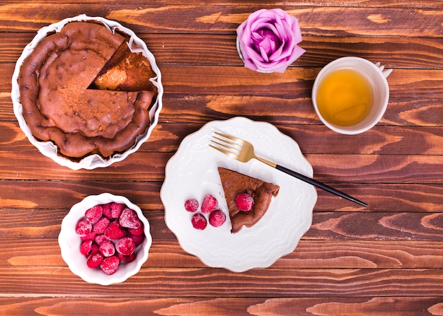 An overhead view of rose; herbal tea; cake slice and raspberry on wooden textured background