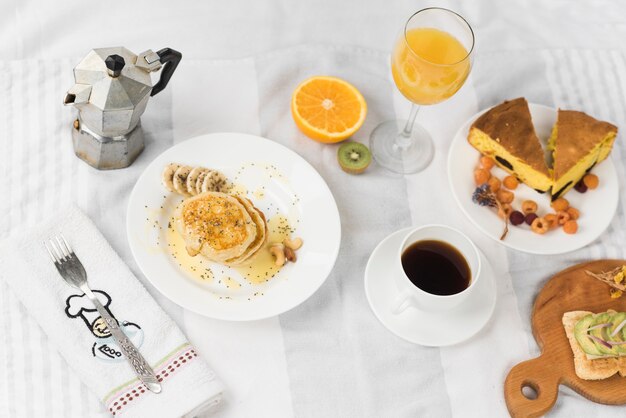 An overhead view of sandwich; pancake; juice; fruits; coffee and cake slice on tablecloth