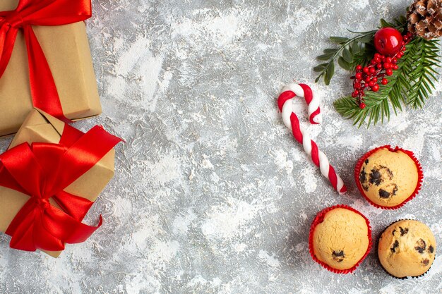 Вид сверху небольших кексов, конфет и аксессуаров для украшения еловых веток и подарков с красной лентой на ледяной поверхности