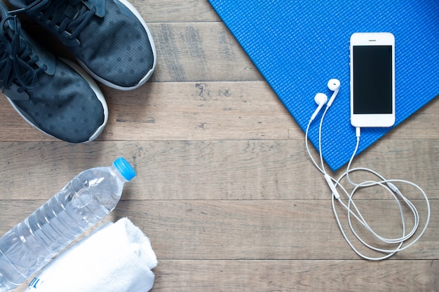 Overhead view of smartphone with earphone on blue yoga mat with black sneaker, bottle of water and towel. Fitness and workout concept with copy space