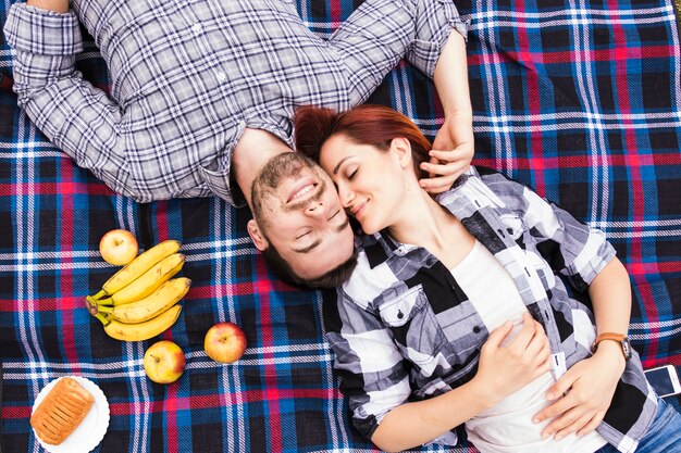An overhead view of smiling romantic young couple lying on blanket with fruits and puff pastry