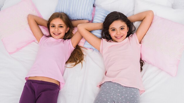 An overhead view of smiling two girls lying on bed looking to camera