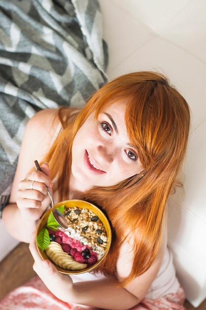 An overhead view of smiling young woman holding bowl of oatmeals with berries