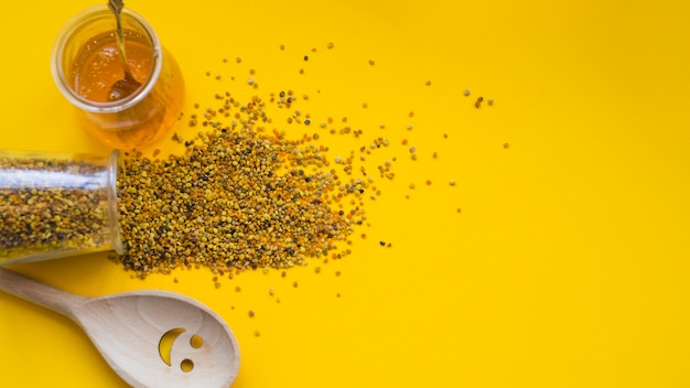 An overhead view of spilled bee pollens; honey pot and smiley wooden spoon on yellow backdrop