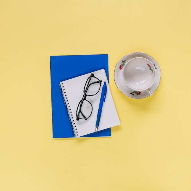 Overhead view of spiral notepad; cup and eyeglasses on yellow surface
