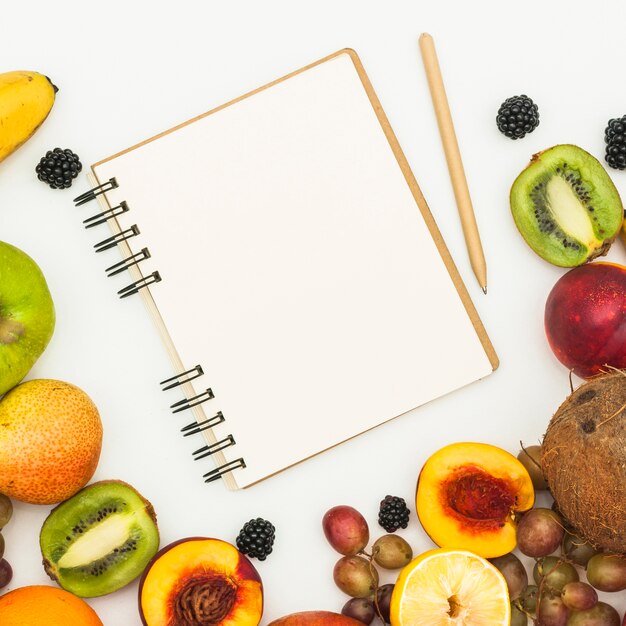 An overhead view of spiral notepad; pencil and various fruits on white backdrop