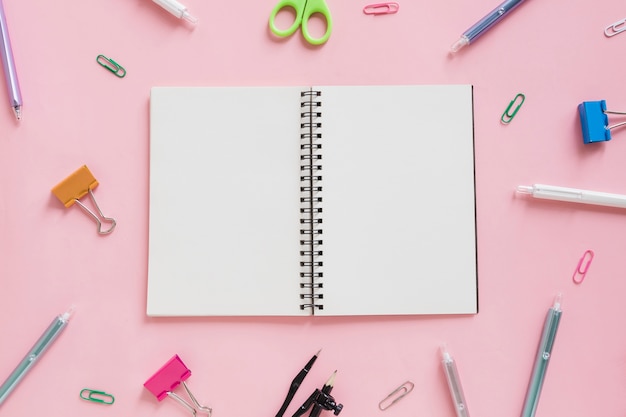 Overhead view of spiral notepad surrounded by various stationeries on pink backdrop