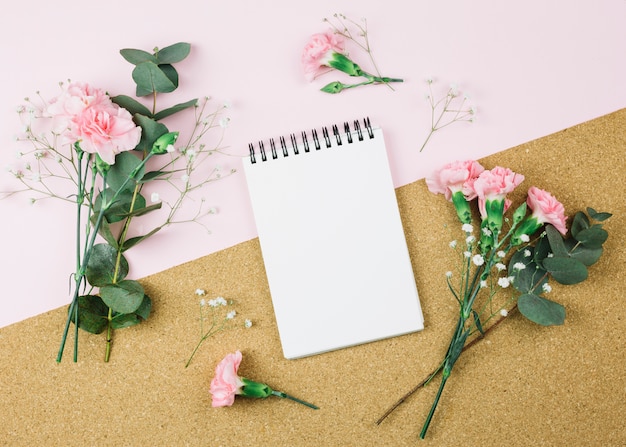 An overhead view of spiral notepad surrounded with gypsophila and carnation flowers on dual pink and cardboard background