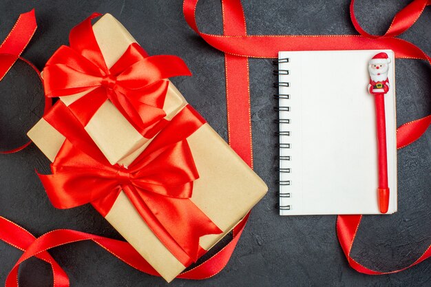 Overhead view of stacked beautiful gifts with red ribbon and notebook with pen on dark background