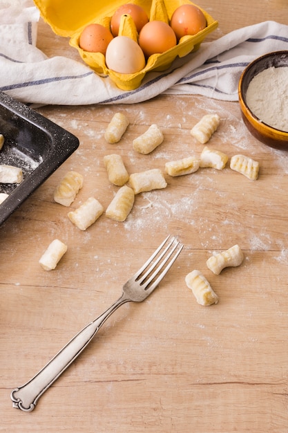 Free photo an overhead view of stainless steel fork with pasta gnocchi dough; eggs and flour on wooden table