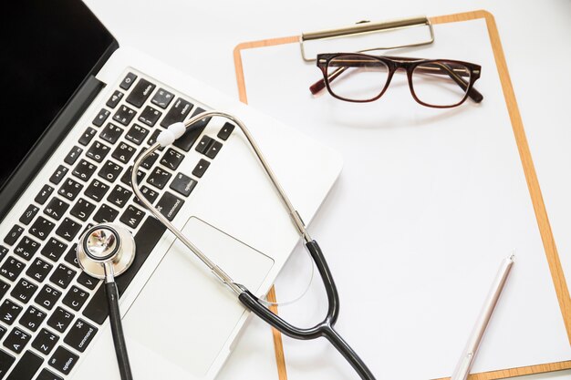 Overhead view of stethoscope on an open laptop with clipboard and eyeglasses on white background