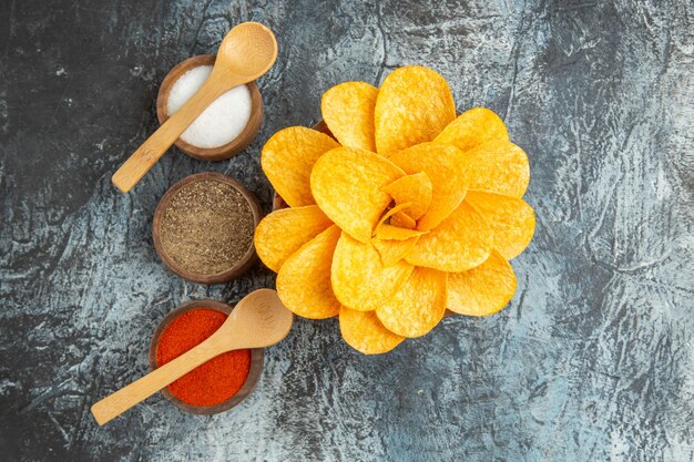 Overhead view of tasty potato chips decorated like flower shaped different spices with spoons on them on gray table